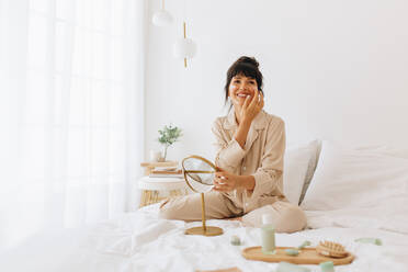 Glückliche Frau im Nachthemd auf dem Bett sitzend und Gesichtscreme auftragend. Frau benutzt Schönheitsprodukte zu Hause. - JLPSF04415