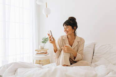 Lächelnde Frau sitzt auf dem Bett und benutzt ihr Handy. Frau im Nachtanzug sitzt zu Hause auf dem Bett und entspannt sich, während sie ihr Handy betrachtet. - JLPSF04432