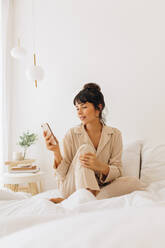 Junge Frau im Nachtanzug sitzt zu Hause auf dem Bett und schaut aufs Handy. Frau entspannt sich zu Hause auf dem Bett und hält ein Handy. - JLPSF04434