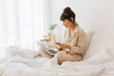 Seitenansicht einer Frau, die auf dem Bett sitzt und einen Laptop benutzt. Eine Frau, die eine Online-Transaktion mit einer Kreditkarte durchführt, sitzt zu Hause. - JLPSF04435