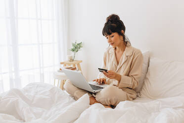 Frau im Nachtanzug sitzt auf dem Bett und benutzt eine Kreditkarte, um eine Online-Transaktion durchzuführen. Frau beim Online-Shopping sitzt auf dem Bett zu Hause. - JLPSF04436