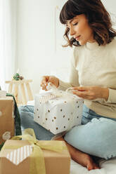 Young woman sitting on bed at home packing christmas presents. Close up of woman tying ribbon on gift box. - JLPSF04440