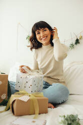 Porträt einer lächelnden Frau, die auf einem Bett mit Geschenkboxen sitzt. Frau feiert Weihnachten zu Hause und macht Weihnachtsgeschenke. - JLPSF04442