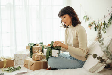 Seitenansicht einer Frau, die zu Hause auf dem Bett sitzt und Weihnachtsgeschenke verpackt. Frau feiert Weihnachten zu Hause. - JLPSF04443