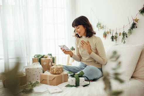 Frau schaut auf Weihnachtsbrief. Frau sitzt auf Bett zu Hause mit Weihnachtsgeschenken und Post. - JLPSF04445