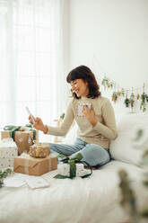 Glückliche Frau sitzt zu Hause mit Geschenk-Boxen und feiern Weihnachten. Frau tut ein Video-Anruf mit Familie und Freunden auf dem Handy. - JLPSF04450