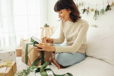 Glückliche Frau schreibt einen Brief an Familie und Freunde anlässlich von Weihnachten. Frau sitzt auf dem Bett mit Weihnachtsgeschenken. - JLPSF04451