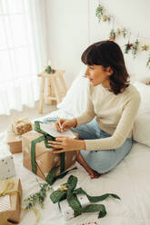 Woman sitting on bed at home with christmas presents and writing letter. Woman celebrating christmas at home with decorations and gifts. - JLPSF04453