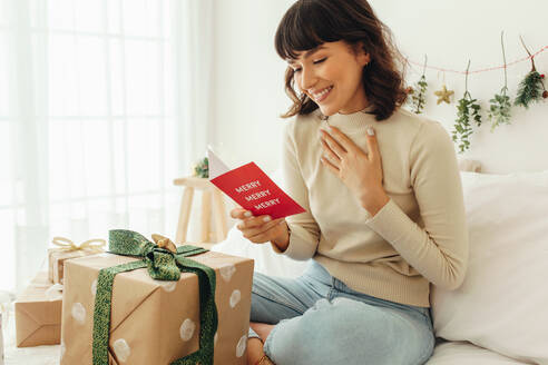Close up of woman sitting on bed with christmas present. Smiling woman holding a christmas card. - JLPSF04455