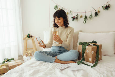 Fröhliche Frau auf dem Bett sitzend mit einem Glas Wein in der Hand und einem Videoanruf auf dem Handy. Frau feiert Weihnachten zu Hause mit Wein und Geschenken. - JLPSF04471