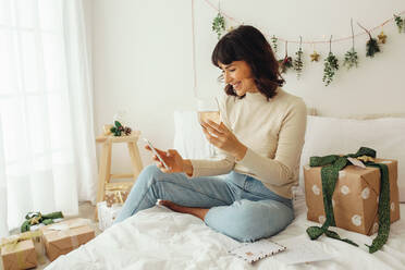 Frau sitzt mit einem Weinglas auf dem Bett und schaut auf ihr Handy. Frau sitzt zu Hause und ist weihnachtlich dekoriert. - JLPSF04472