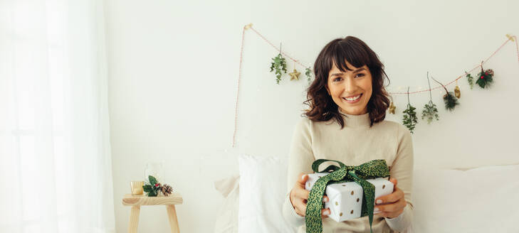 Lächelnde Frau, die ein Weihnachtsgeschenk in der Hand hält, mit Dekoration im Hintergrund. - JLPSF04477