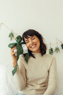 Nahaufnahme einer lächelnden Frau, die ein Weihnachtsgeschenk hält. Glückliche Frau sitzt mit einem Weihnachtsgeschenk mit Dekoration im Hintergrund. - JLPSF04478