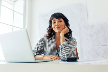 Lächelnde Architektin bei der Arbeit am Laptop im Büro. Unternehmerin sitzt am Arbeitsplatz mit Architekturzeichnungen im Hintergrund. - JLPSF04493