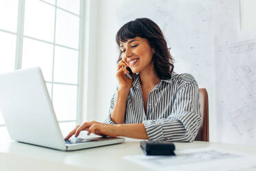 Lächelnde Architektin, die im Büro am Laptop arbeitet, während sie über das Mobiltelefon spricht. Weibliche Unternehmerin sitzt am Arbeitsplatz mit Architekturzeichnungen im Hintergrund. - JLPSF04495