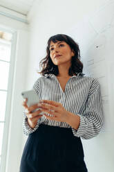 Low angle shot of woman entrepreneur standing in office holding mobile phone. Portrait of businesswoman standing in office. - JLPSF04506