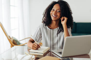 Afrikanische junge Frau, die von zu Hause aus arbeitet. Die Frau lächelt in die Kamera, während sie mit ihrem Tagebuch und ihrem Laptop am Tisch sitzt. - JLPSF04515