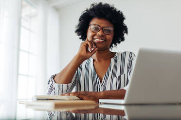 Happy African woman sitting at desk at home. Woman smiling at camera while working from home. - JLPSF04520