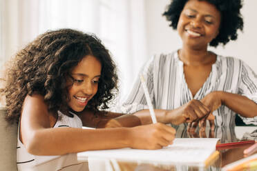 Girl writing her homework while grandmother observes. Small girl doing homework with grandma sitting by - JLPSF04540