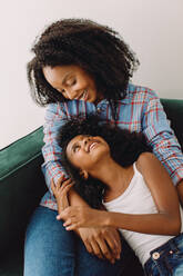 Fürsorgliche Mutter mit ihrer Tochter zu Hause. Afrikanische Frau betrachtet ihre Tochter auf ihrem Schoß, während sie auf dem Sofa sitzt. - JLPSF04573