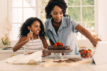 Mutter und Tochter, die einen Kuchen backen, während sie durch ein Online-Tutorial von einem digitalen Tablet zu Hause lernen. Frau schaut auf ihren Tablet-PC, während sie mit ihrer Tochter in der Küche einen Kuchen zubereitet. - JLPSF04604