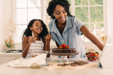 Lächelndes Mädchen mit Mutter, die auf ein digitales Tablet schaut, während sie in der Küche einen Kuchen zubereiten. Mutter und Tochter folgen einem Online-Kochkurs zu Hause. - JLPSF04605