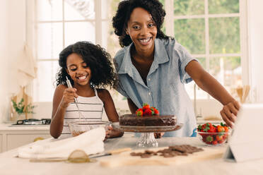Afrikanische Frau mit Tochter, die zu Hause einen Geburtstag feiern. Lächelnde Mutter und Tochter, die online mit einem Tablet-PC lernen, einen Kuchen zu backen. - JLPSF04606