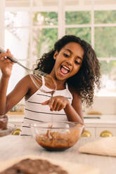 Girl licking chocolate cream while baking in kitchen. Cute children making batter for baking. - JLPSF04616