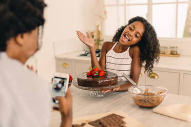 Glückliches junges Mädchen steht in der Küche mit Kuchen und wird von ihrer Mutter fotografiert. Tochter mit Schokoladenkuchen zu Hause mit ihrer Mutter, die sie mit dem Handy fotografiert. - JLPSF04620