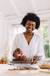 Afrikanische Seniorin beim Verzieren von Schokoladenkuchen in der Küche. Frau beim Verzieren von Kuchen mit Erdbeeren. - JLPSF04628