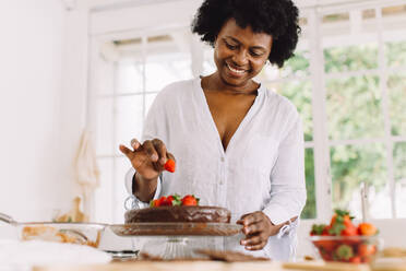 Glückliche ältere Frau, die Erdbeeren auf einen Schokoladenkuchen in der Küche legt. Afrikanische Frau bereitet zu Hause leckeren Kuchen zu. - JLPSF04629