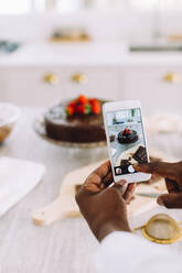 Close-up of a female hands holding mobile phone and taking pictures of cake on kitchen counter. Woman photographing freshly made cake at home. - JLPSF04633