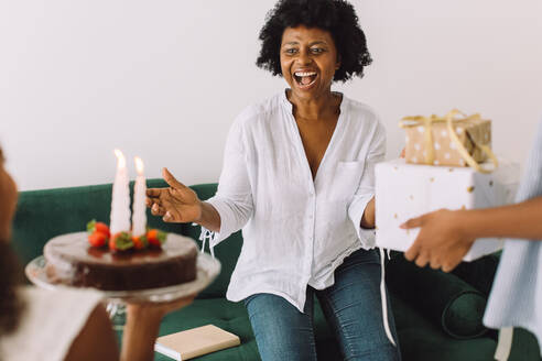 Reife Frau, die von ihrer Tochter überrascht wird, die ihr Geburtstagskuchen und Geschenke bringt. Mädchen feiern den Geburtstag ihrer Mutter zu Hause. - JLPSF04634