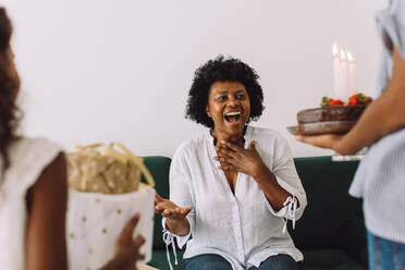 Reife Frau wird von ihren Töchtern zu Hause mit Geburtstagskuchen und Geschenken überrascht. Kleine Familie feiert Geburtstag zu Hause. - JLPSF04636