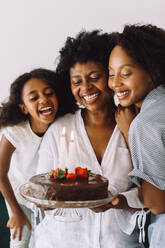 Mature woman holding her birthday cake with her daughters standing by. Woman celebrating her birthday at home with her daughters. - JLPSF04640