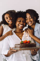 Mädchen, die zu Hause den Geburtstag ihrer Mutter feiern. Eine reife Frau, die ihren Geburtstagskuchen hält, während ihre Töchter lächeln. - JLPSF04642