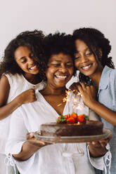 Cheerful family of three celebrating birthday at home. Grandmother holding her birthday cake with daughter and granddaughter standing by. - JLPSF04643