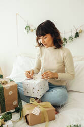 Frau bindet Bänder an Weihnachtsgeschenke auf dem Bett sitzend. Frau trifft Vorbereitungen für das Weihnachtsfest. - JLPSF04665