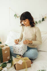 Woman tying ribbons to christmas presents sitting on bed. Woman making preparations for celebrating christmas. - JLPSF04665