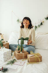 Glückliche Frau sitzt auf dem Bett mit Weihnachtsgeschenken. Junge Frau hält Weihnachtsgeschenk zu Hause. - JLPSF04666