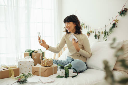 Frau feiert Weihnachten zu Hause im Gespräch über Videoanruf mit Mobiltelefon. Glückliche Frau sitzt auf dem Bett mit Weihnachtsgeschenken und Briefen. - JLPSF04668