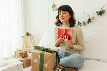 Nahaufnahme einer Frau, die mit Weihnachtsgeschenken auf dem Bett sitzt und mit geschlossenen Augen eine Weihnachtskarte an ihre Brust hält. - JLPSF04669