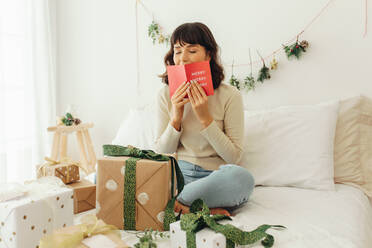 Frau feiert Weihnachten zu Hause sitzend mit Geschenken. Frau sitzt auf dem Bett und küsst eine Weihnachtskarte mit geschlossenen Augen. - JLPSF04670