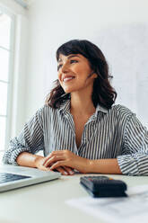 Portrait of businesswoman sitting at her desk in office. Smiling woman at work with laptop and calculator on desk. - JLPSF04694