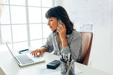 Female entrepreneur managing business talking over mobile phone. Businesswoman working on laptop sitting at her desk. - JLPSF04696