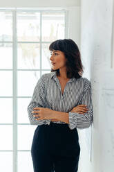 Woman entrepreneur standing in office with arms crossed. Confident businesswoman standing in office and looking away. - JLPSF04701