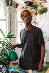 Lächelnder schwuler Mann, der zu Hause einen Blumentopf gießt. Junger schwarzer Mann, der fröhlich in die Kamera schaut, während er sich um seine Topfpflanze kümmert. Junger Mann, der allein steht. - JLPSF04732