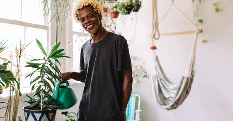 Happy young gay man watering a flower pot indoors at home. Young black man smiling at the camera cheerfully while taking care of his pot plant. Young man standing alone. - JLPSF04733