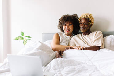 Unbeschwertes schwules Paar, das seine Lieblingssendung auf einem Laptop schaut. Zwei glückliche junge männliche Liebhaber, die fröhlich lächelnd zusammen im Bett sitzen. Junges schwules Paar, das seine Zeit zusammen zu Hause verbringt. - JLPSF04761