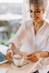 Lächelnde Geschäftsfrau, die eine Kaffeepause macht, während sie in einem Café arbeitet. Reife Geschäftsfrau, die eine Tasse Kaffee trinkt, während sie allein an einem Tisch sitzt. Geschäftsfrau, die an einem Laptop arbeitet. - JLPSF04825